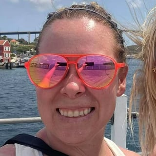 Smiling person wearing bright pink heart-shaped sunglasses on a boat with a waterfront town visible in the background