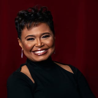 Portrait of a smiling woman with short black hair, wearing a black top with shoulder cutouts and silver earrings against a deep red background